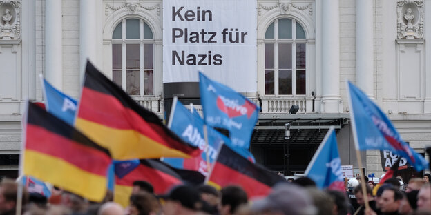 Menschen tragen Deutschland- und AfD-Fahnen, dahinter hängt ein Transparent an einem Gebäude, auf dem „Kein Platz für Nazis“ steht Menschen tragen Deutschland- und AfD-Fahnen, dahinter hängt ein Transparent an einem Gebäude, auf dem „Kein Platz für Nazis“ steht