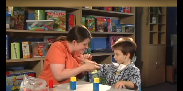 Eine Frau und ein Junge sitzen an einem Tisch, dahinter ein Regal voller Spiele Eine Frau und ein Junge sitzen an einem Tisch, dahinter ein Regal voller Spiele