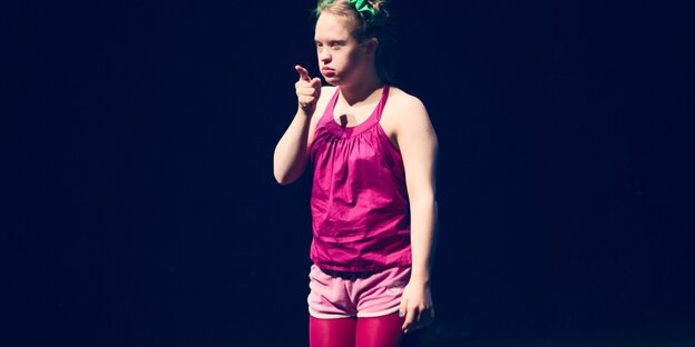 Eine Frau mit rosafarbener und pinker Kleidung steht auf einer Theaterbühne und zeigt mit einem Finger nach vorn. Sie hat das Down-Syndrom Eine Frau mit rosafarbener und pinker Kleidung steht auf einer Theaterbühne und zeigt mit einem Finger nach vorn. Sie hat das Down-Syndrom