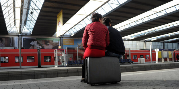 Eine junge Frau in roter Jacke und ein Mann sitzen auf einem Koffer und wartet auf den Zug Eine junge Frau in roter Jacke und ein Mann sitzen auf einem Koffer und wartet auf den Zug