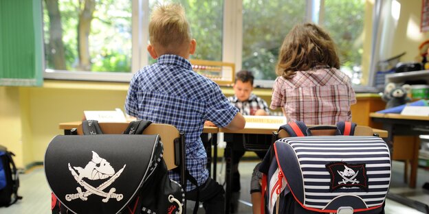 Zwei Grundschüler*innen von hinten, über den Stühlen hängen die Schulranzen, die Kinder brüten über einer Aufgabe Zwei Grundschüler*innen von hinten, über den Stühlen hängen die Schulranzen, die Kinder brüten über einer Aufgabe
