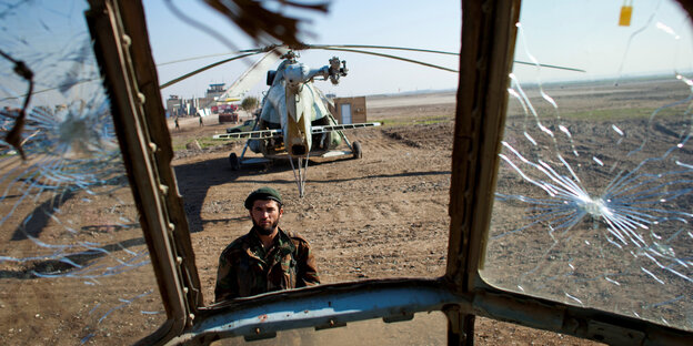 Blick durch ein zersplittertes Cockpit auf einen Soldaten, dahinter ein Hubschrauber
