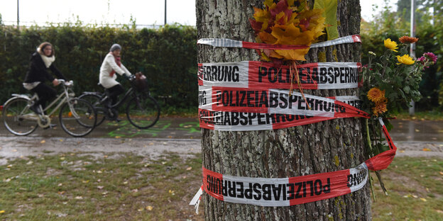 Um einen Baum ist Absperrband der Polizei gewunden, in das Blumen und Blätter gesteckt wurden Um einen Baum ist Absperrband der Polizei gewunden, in das Blumen und Blätter gesteckt wurden
