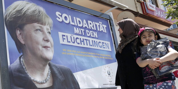 Auf eine Plakat ist Angela Merkel und der Satz „Solidarität mit Flüchtlingen“ zu sehen, davor steht eine Frau und ein Kind Auf eine Plakat ist Angela Merkel und der Satz „Solidarität mit Flüchtlingen“ zu sehen, davor steht eine Frau und ein Kind