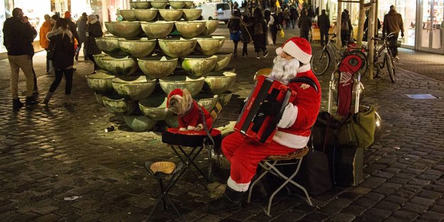 Ein Mann im Weihnachtsmannkostüm spielt Akkordeon auf einem Platz. Neben ihm sitzt ein Hund Ein Mann im Weihnachtsmannkostüm spielt Akkordeon auf einem Platz. Neben ihm sitzt ein Hund