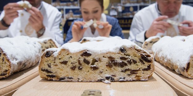 Ein angeschnittener Christstollen, im Hintergrund Menschen, die ihn probieren Ein angeschnittener Christstollen, im Hintergrund Menschen, die ihn probieren