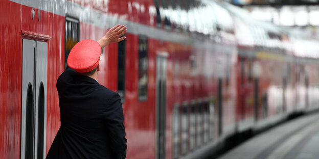 Ein Bahnmitarbeiter steht neben einem roten Zug und winkt mit der linken Hand Ein Bahnmitarbeiter steht neben einem roten Zug und winkt mit der linken Hand