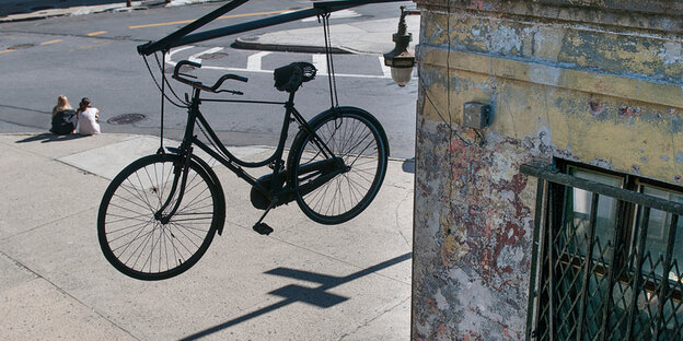 Ein Fahrrad hängt an einer Fassade. An einer Straßenecke sitzen zwei Personen. Ein Fahrrad hängt an einer Fassade. An einer Straßenecke sitzen zwei Personen.