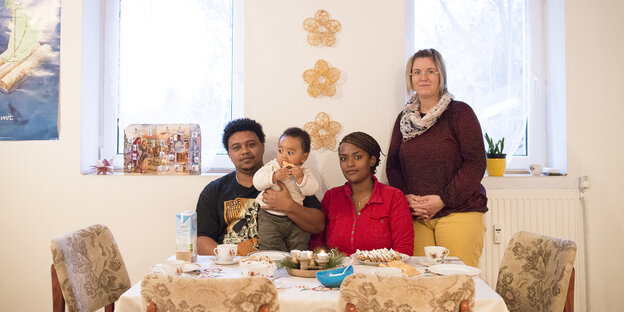 Vater, Mutter und Kind sitzen an einem Tisch mit Christstollen und Kerzen, daneben steht eine Frau Vater, Mutter und Kind sitzen an einem Tisch mit Christstollen und Kerzen, daneben steht eine Frau