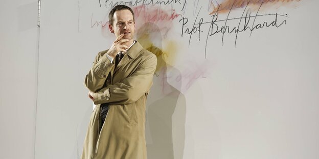 Ein Mann in einem grau-grünen Mantel steht an einer weißen Wand gelehnt Ein Mann in einem grau-grünen Mantel steht an einer weißen Wand gelehnt