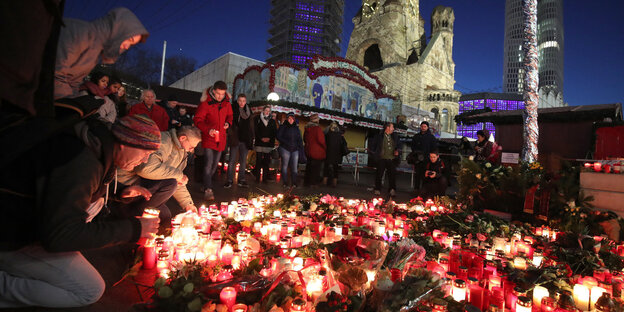 Viele Blumen und Kerzen, daneben sitzen und stehen Menschen Viele Blumen und Kerzen, daneben sitzen und stehen Menschen