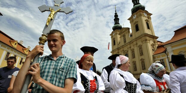 Ein junger Mann trägt ein Kreuz, an dem eine Jesusfigur hängt, dahinter läufen Frauen in Trachten Ein junger Mann trägt ein Kreuz, an dem eine Jesusfigur hängt, dahinter läufen Frauen in Trachten
