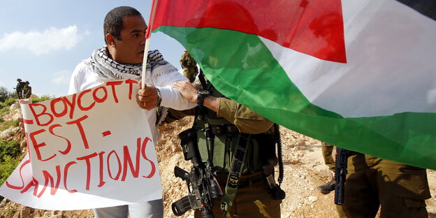 Ein Aktivist hält eine palästinensischen Flagge und ein Schild mit „Boycott, Divestment and Sanctions“ in der Hand Ein Aktivist hält eine palästinensischen Flagge und ein Schild mit „Boycott, Divestment and Sanctions“ in der Hand