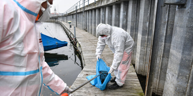 Zwei weiß vermummte Gestalten stehen auf einem Steg am Wasser packen etwas in einen blauen Abfallbeutel Zwei weiß vermummte Gestalten stehen auf einem Steg am Wasser packen etwas in einen blauen Abfallbeutel