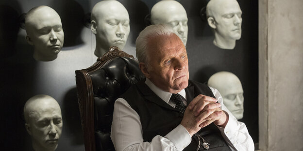 Anthony Hopkins sitzt, in seiner Rolle in "Westworld", nachdenklich vor einer Wand mit weißen Masken Anthony Hopkins sitzt, in seiner Rolle in "Westworld", nachdenklich vor einer Wand mit weißen Masken