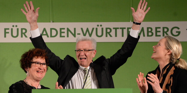 Ein Mann in der Mitte steckt freudig beide Hände nach oben, zwei Frauen stehen rechts und links neben ihm Ein Mann in der Mitte steckt freudig beide Hände nach oben, zwei Frauen stehen rechts und links neben ihm