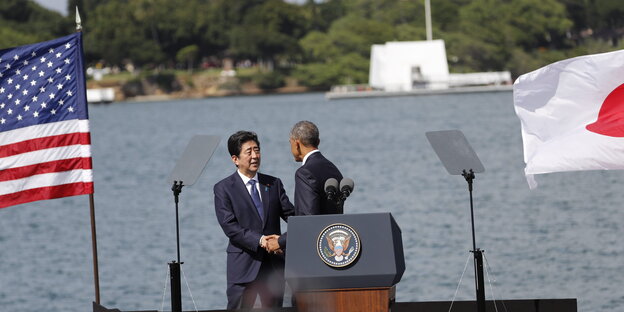 Zwei Männer schütteln sich vor einem Podest die Hände, rechts die japanische, links die US-amerikanische Flagge. Zwei Männer schütteln sich vor einem Podest die Hände, rechts die japanische, links die US-amerikanische Flagge.
