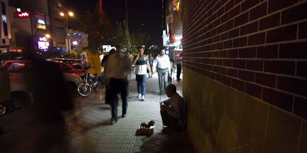 Es ist Nacht, eine belebte Straße, an einer Hauswand sitzt ein Mann Es ist Nacht, eine belebte Straße, an einer Hauswand sitzt ein Mann