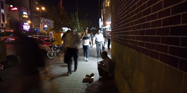 Es ist Nacht, eine belebte Straße, an einer Hauswand sitzt ein Mann