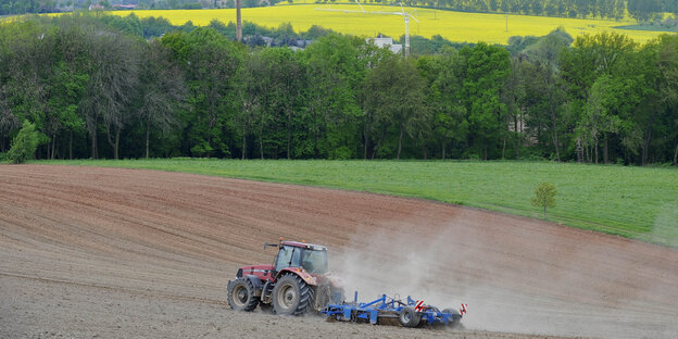 Ein Traktor ackert ein Feld um. Im Hintergrund sind Rapsfelder und Wald zu sehen.