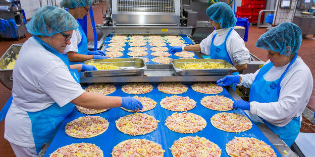 Am Fließband stehen zwei Damen und belegen Tiefkühlpizzen. Am Fließband stehen zwei Damen und belegen Tiefkühlpizzen.