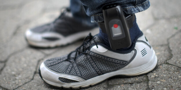 Zwischen Turnschuhen und Jeans eines Mannes ist eine elektronische Fußfessel angebracht Zwischen Turnschuhen und Jeans eines Mannes ist eine elektronische Fußfessel angebracht