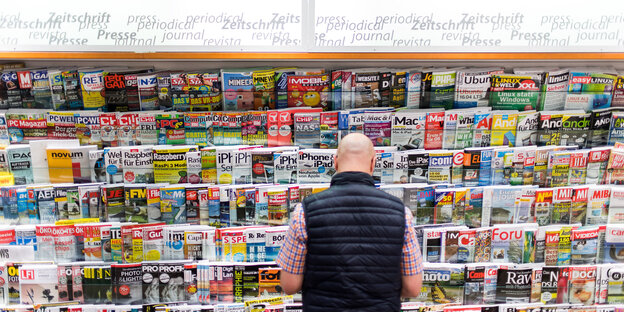 Ein Mann in Weste steht mit dem Rücken zum Bild vor einem Zeitschriftenständer. Ein Mann in Weste steht mit dem Rücken zum Bild vor einem Zeitschriftenständer.