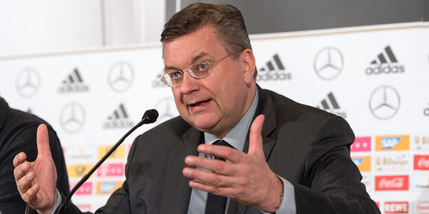 Ein Mann mit Brille und in Nazug sitzt an einem Mikrofon und gestikuliert beim Sprechen Ein Mann mit Brille und in Nazug sitzt an einem Mikrofon und gestikuliert beim Sprechen