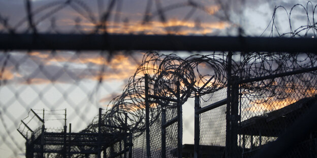 Stacheldrahtzaun rankt sich überall um Guantanamo, dahinter ein rosig-blauer Abendhimmel Stacheldrahtzaun rankt sich überall um Guantanamo, dahinter ein rosig-blauer Abendhimmel