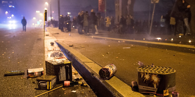 Auf einer Straße liegen Reste von Feuerwerk, dahinter auf dem Gehweg stehen Menschen. Es ist Nacht Auf einer Straße liegen Reste von Feuerwerk, dahinter auf dem Gehweg stehen Menschen. Es ist Nacht