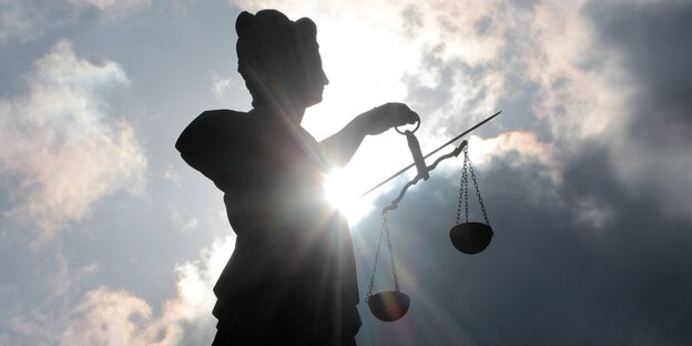 Silhouette einer Justitia-Statue mit Schwert und Waagschalen vor dunklen Wolken Silhouette einer Justitia-Statue mit Schwert und Waagschalen vor dunklen Wolken