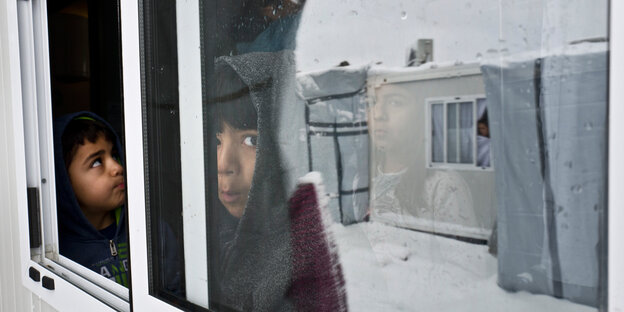 Kinder blicken aus dem Fenster einer Unterkunft, im Fenster spiegeln sich verschneite Wohncontainer Kinder blicken aus dem Fenster einer Unterkunft, im Fenster spiegeln sich verschneite Wohncontainer