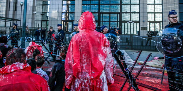 Menschen, die mit Kunstblut getränkte Overalls tragen, stehen hinter Stacheldrahtzaun vor einem Gebäude, das von Soldaten geschützt wird