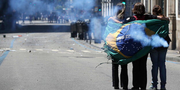 Drei Menschen haben sich in eine brasilianische Fahne eingehüllt, um sie herum weht Rauch Drei Menschen haben sich in eine brasilianische Fahne eingehüllt, um sie herum weht Rauch