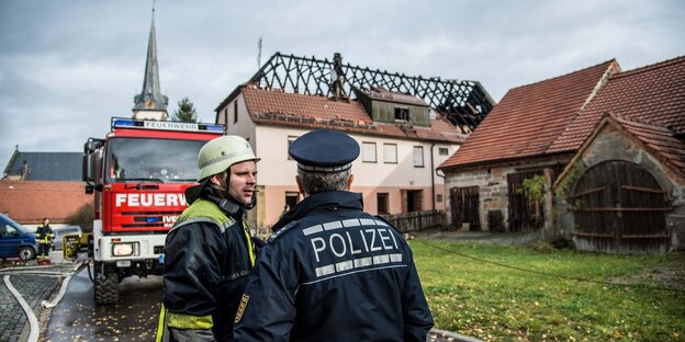 Neben einem Feuerwehrauto stehen ein Feuerwehrmann und ein Polizist. Sie blicken auf eine altes Haus, das Brandspuren aufweist