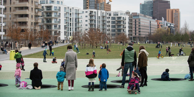 Auf einem grünen Boden stehen Erwachsene und Kinder in bunter Kleidung, dahinter stehen Mehrfamilienhäuser Auf einem grünen Boden stehen Erwachsene und Kinder in bunter Kleidung, dahinter stehen Mehrfamilienhäuser