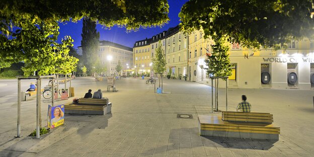 Hellerleuchteter städtischer Platz bei Dunkelheit. Von Bäumen umrahmt, Leute sitzen auf Bänken Hellerleuchteter städtischer Platz bei Dunkelheit. Von Bäumen umrahmt, Leute sitzen auf Bänken