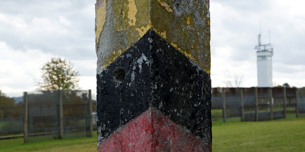 Ein schwarz-rot-gold bemalter Pfahl vor einem Zaun. Dahinter ein Wachturm