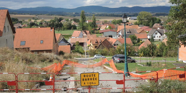 Eine gesperrte Straße, dahinter ein Dorf, am Horizont Berge Eine gesperrte Straße, dahinter ein Dorf, am Horizont Berge