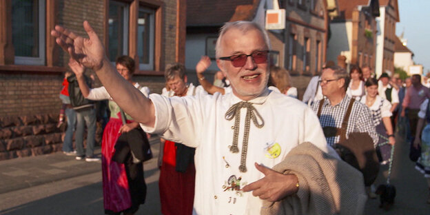 Ein Straßenumzug in einer ländlichen Straße, ein Mann winkt, er hat ein traditionelles Hemd an