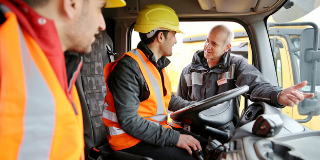 Zwei Männer in orangen Westen im Fahrerhaus eines LKW, ein weiterer steht in der Tür und redet Zwei Männer in orangen Westen im Fahrerhaus eines LKW, ein weiterer steht in der Tür und redet