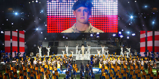 Auf einer Riesenleinwand ist ein Soldat zu sehen, darunter tanzen Cheerleader Auf einer Riesenleinwand ist ein Soldat zu sehen, darunter tanzen Cheerleader