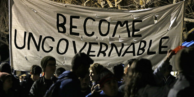 Menschen stehen bei schwacher Beleuchtung beieinander. Auf einem Banner steht: Become Ungovernable Menschen stehen bei schwacher Beleuchtung beieinander. Auf einem Banner steht: Become Ungovernable