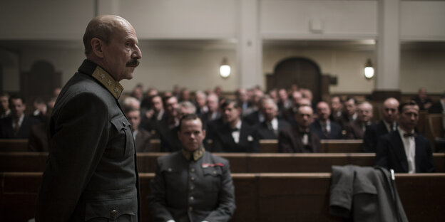 Ein Mann in Uniform steht vor einer Menschen, die in einem Saal auf Bänken sitzen