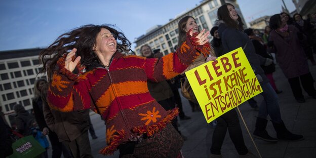 Frau tanzt neben Plakat mit der Aufschrift "Liebe ist keine Entschuldigung", dahinter Menschenmenge