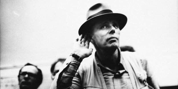 Joseph Beuys in Weste und mit typischem Hut hält sich süffisant die Hand ans rechte Ohr Joseph Beuys in Weste und mit typischem Hut hält sich süffisant die Hand ans rechte Ohr
