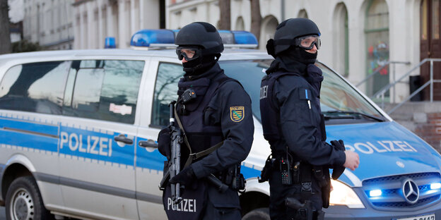 Zwei Polizisten in voller Schutzmontur stehen Rücken an Rücken vor einem Polizeiauto Zwei Polizisten in voller Schutzmontur stehen Rücken an Rücken vor einem Polizeiauto