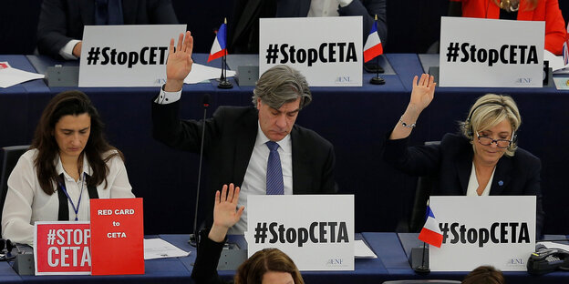 Mehrere Menschen sitzen mit "Sotp Ceta"-Schildern an Tischen und heben die Hände Mehrere Menschen sitzen mit "Sotp Ceta"-Schildern an Tischen und heben die Hände