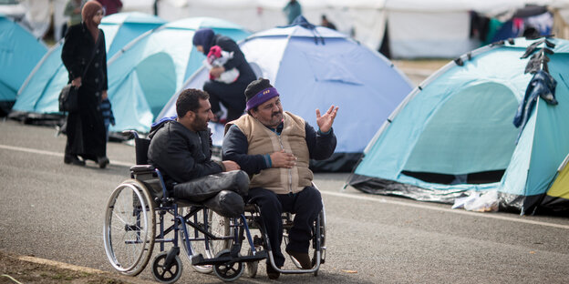 Zwei Männer in Rollstühlen unterhalten sich miteinander, im Hintergrund eine Zeltstadt und weitere Menschen Zwei Männer in Rollstühlen unterhalten sich miteinander, im Hintergrund eine Zeltstadt und weitere Menschen