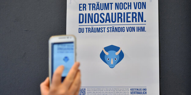 Plakat mit Aufdruck "Er träumt noch von Dinosauriern. Du träumst ständig von ihm“, davor eine Kinderhand Plakat mit Aufdruck "Er träumt noch von Dinosauriern. Du träumst ständig von ihm“, davor eine Kinderhand